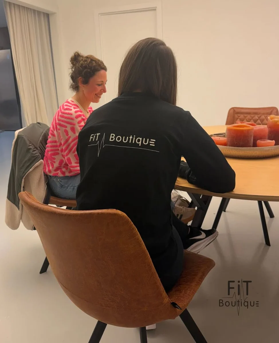 Persoon in zwarte Fit Boutique-trui zit aan tafel in gesprek met een glimlachende vrouw in roze trui